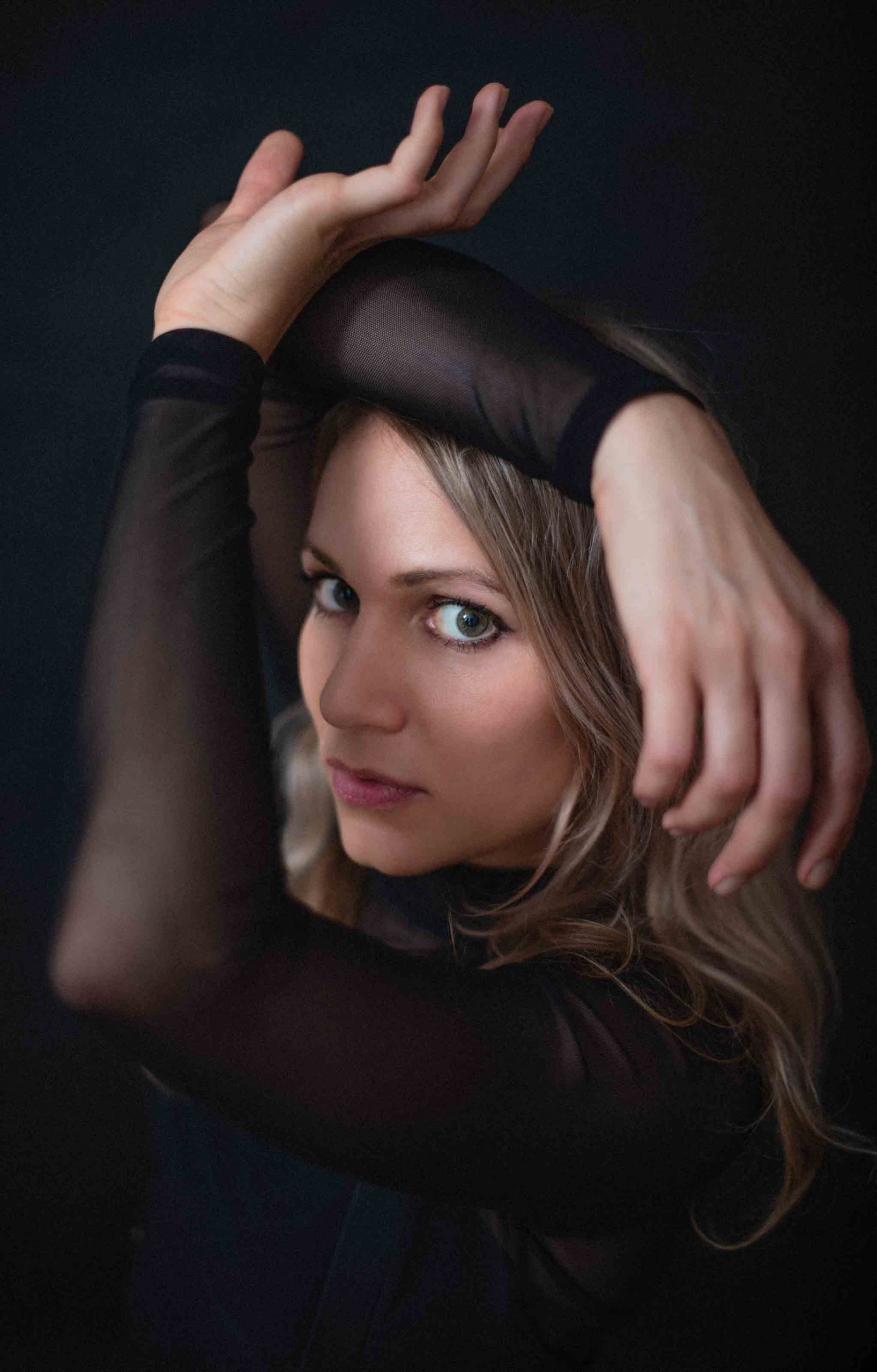 Retrato de la pintora Anne Elizza con los brazos sobre la cabeza sobre fondo oscuro