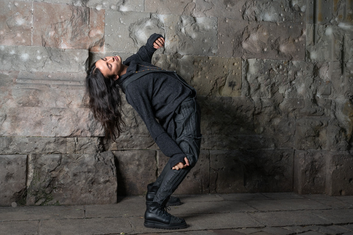 Modelo con conjunto negro arqueando el cuerpo ante un muro de piedra