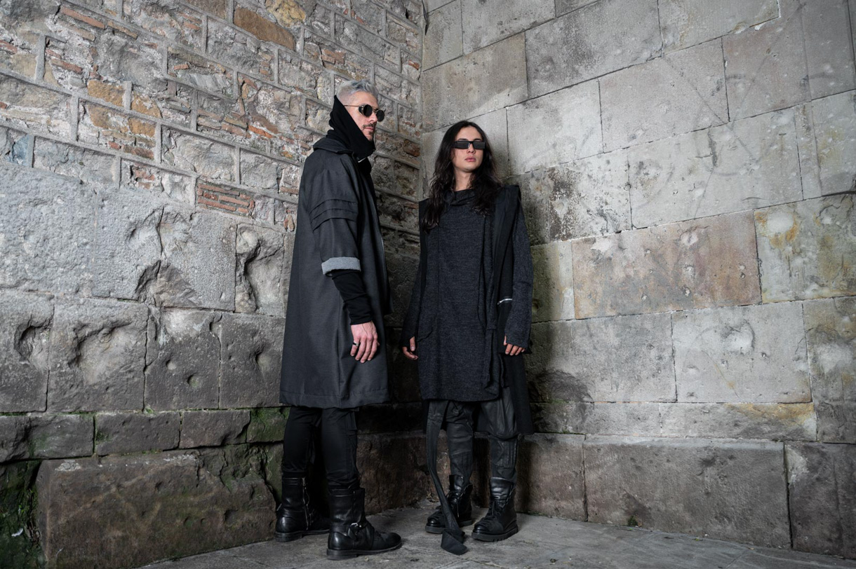 Dos modelos vestidos de negro junto a un muro de piedra