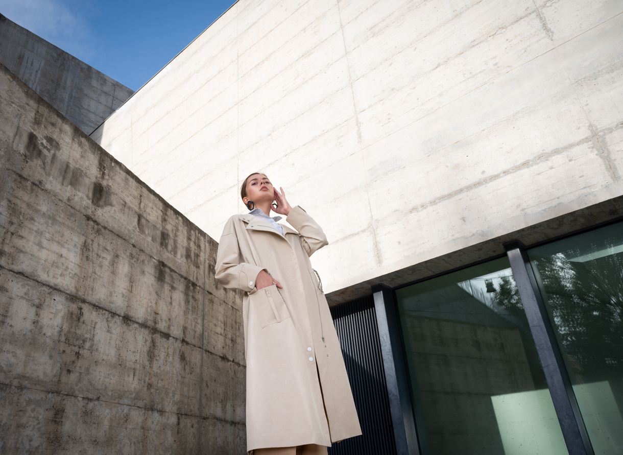 Modelo con abrigo beige fotografiada a contrapicado junto a un muro de hormigón