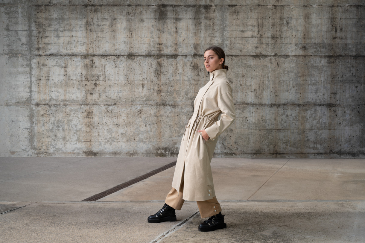 Modelo con gabardina beige caminando frente a un muro de hormigón