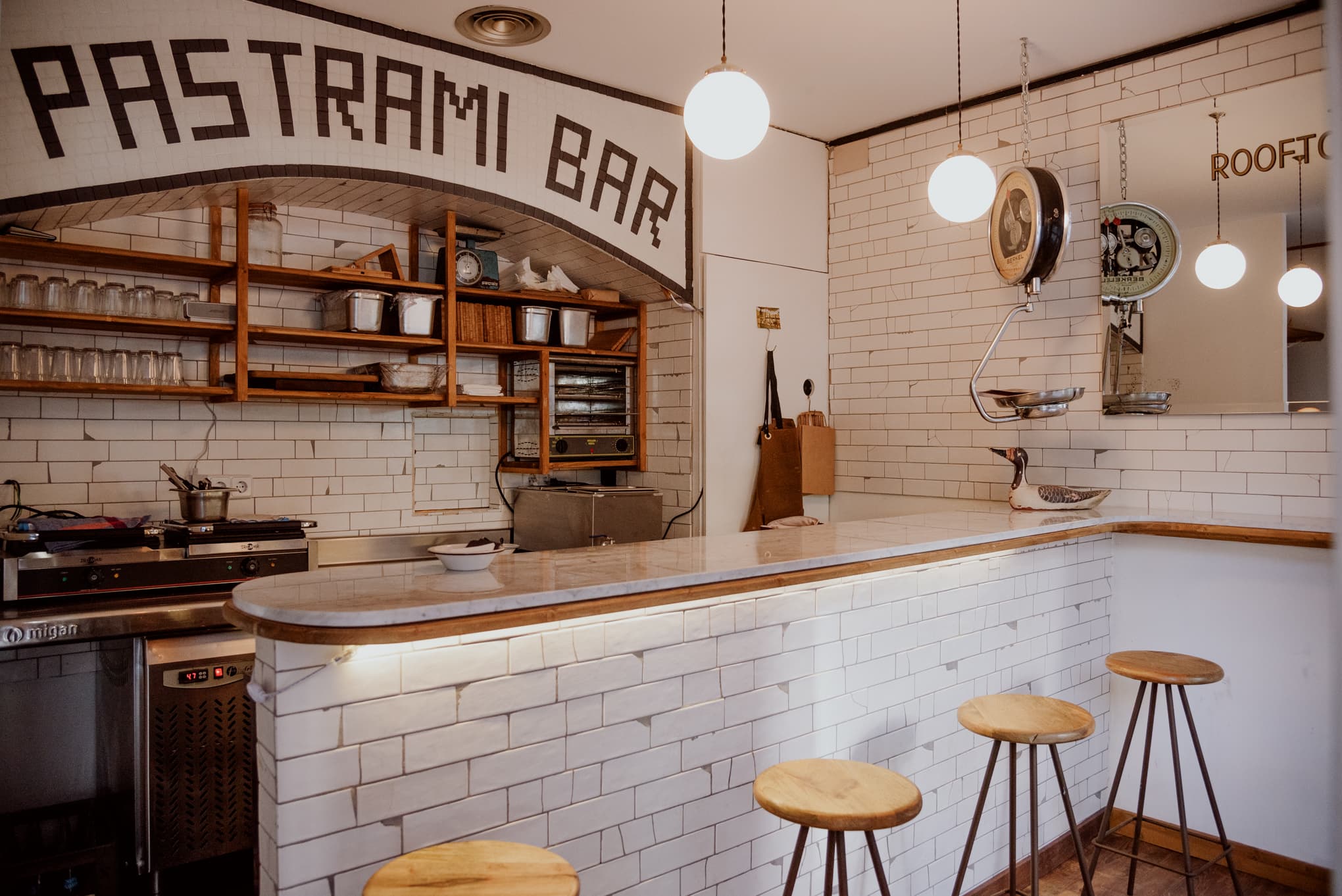 Barra del Pastrami Bar con azulejo blanco, taburetes de madera y lámparas esféricas