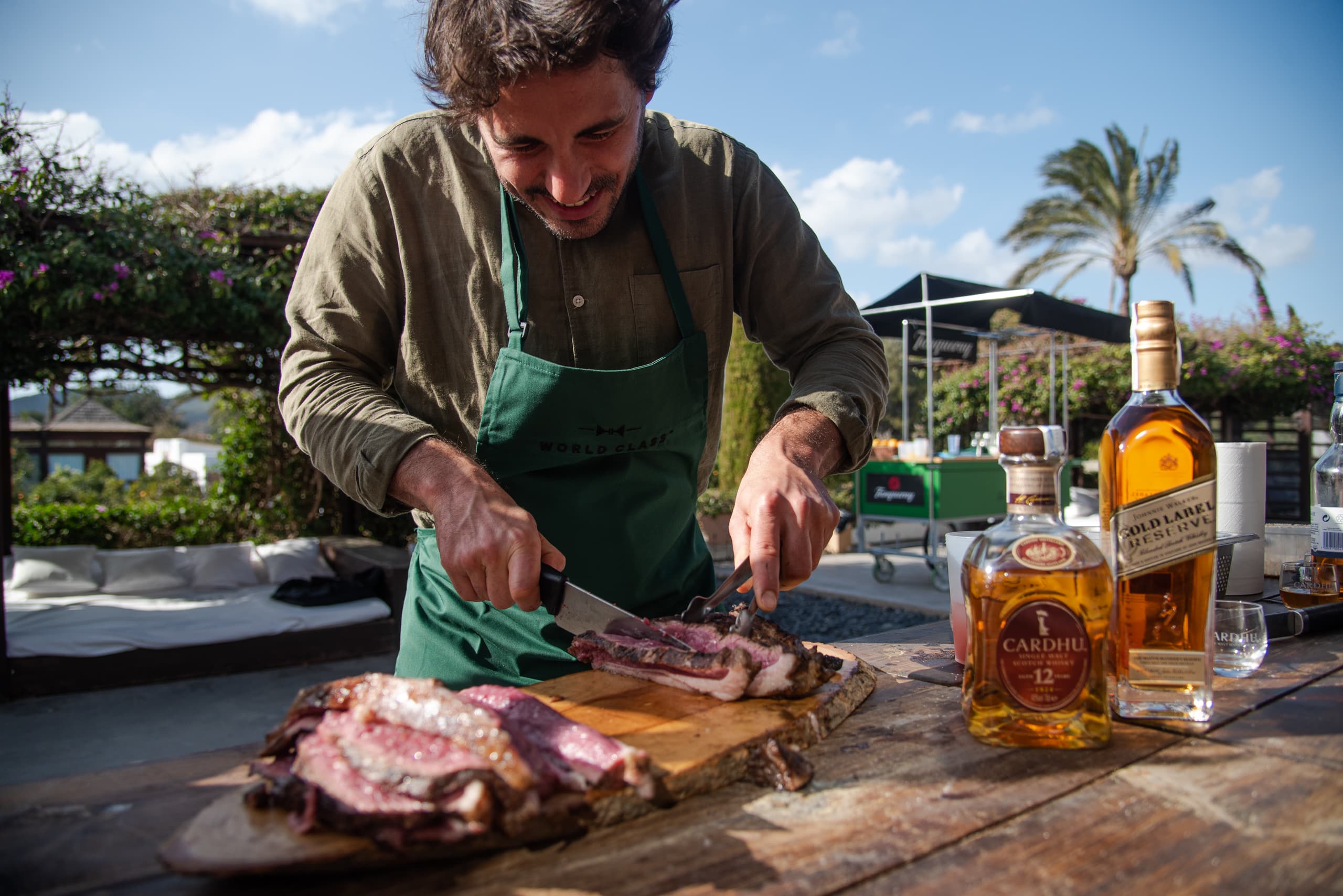 Cocinero cortando carne junto a botellas de whisky en una mesa al aire libre