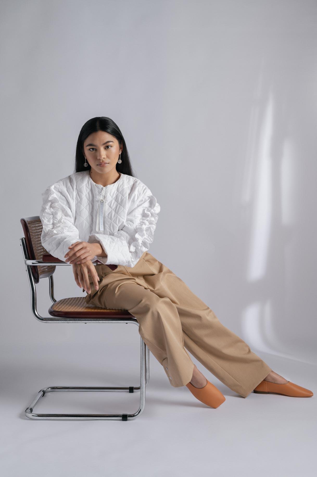 Modelo sentada en una silla con blusa blanca y pantalón camel